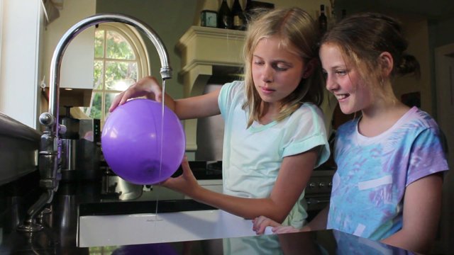 Two girls placing blown up balloon under tap of running water.