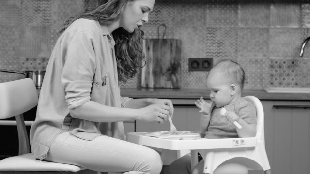 Mother sits feeding baby in highchair.